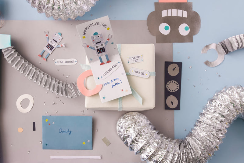 Flat lay of a Fathers Day robot card craft with a wrapped gift, robot cutouts, and metallic streamers.