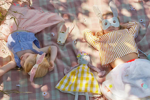 Two children wearing bunny ears lie on a pastel Easter picnic blanket with a plush bunny toy.