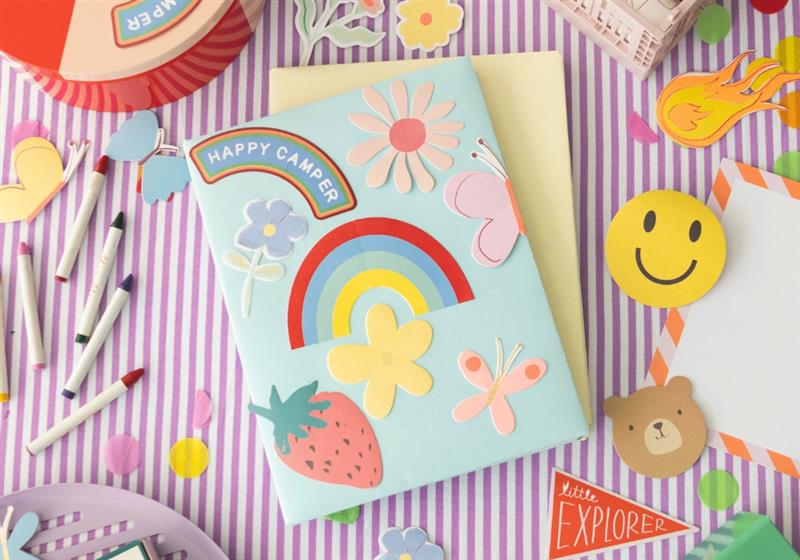 Colorful back to school flat lay with a pastel blue Happy Camper notebook, rainbow motif and markers on a striped surface