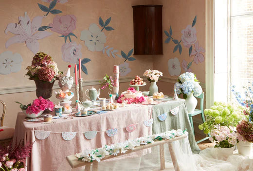 Pastel tea party setup with flowers, cupcakes, and teaware on a long table against a floral wall.