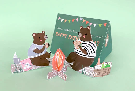Two brown bears sit by a small campfire as a green Father's Day card with bunting stands behind them.