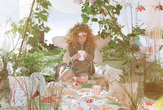 Child dressed as a fairy with wings sits drinking from a cup under leafy decor at a woodland fairy party setup.