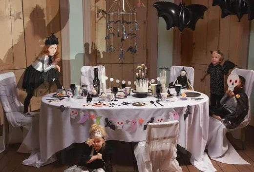 Halloween party scene with a round white-draped table black bat balloons and costumed children.