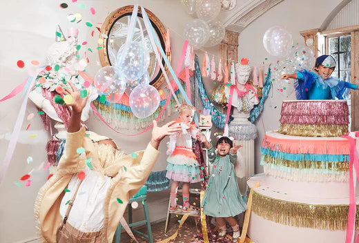 People in festive costumes celebrate Mardi Gras with confetti balloons and a tall multi tier cake in a decorated room.