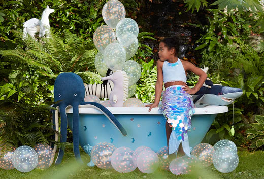 Young girl in a blue mermaid tail costume sits on the edge of a blue tub outdoors, surrounded by balloons.