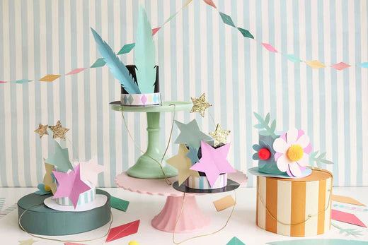 Various circus hats made from coloured card and in different designs such as stars and flowers are displayed against a striped blue background and below a pastel diamond-shaped garland.