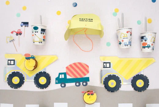 Several construction truck plates are placed on a table with construction-themed cups and a card hard hat headpiece.