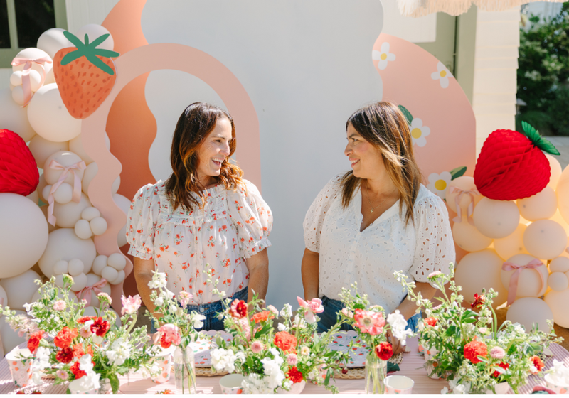 Two women chat at a strawberry themed party with pink peach decor and balloon backdrop