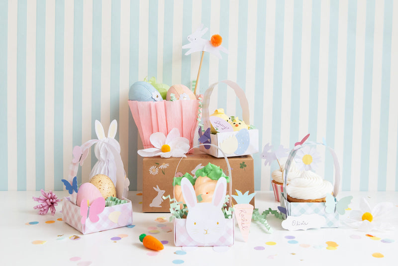 Easter craft scene with pastel baskets, paper eggs, bunny toppers, flowers and confetti on a striped backdrop.