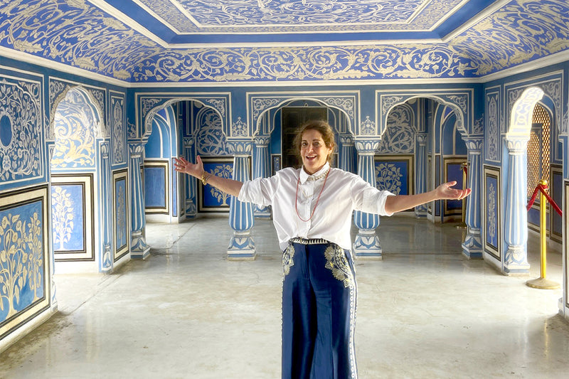 Woman in white blouse stands with arms wide inside a blue ornate Jaipur room with arches and columns.