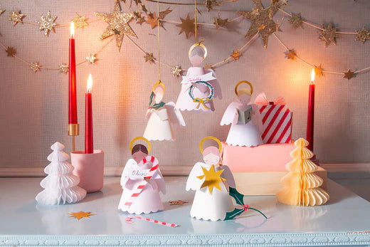 Paper angel ornaments stand on the table and some hang from the ceiling along with a star garland. Honeycomb trees are placed beside lighted red taper candles.