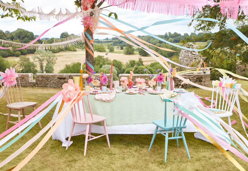 Pastel outdoor dinner party setup with a long table, ribbons overhead, and pink flowers in a countryside garden.