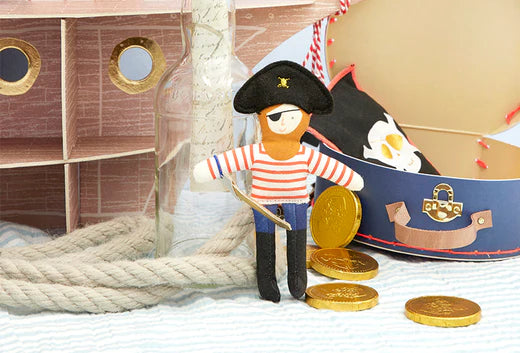 Handmade pirate figure in a striped shirt stands beside a toy ship with coins and a rope on a wooden surface.
