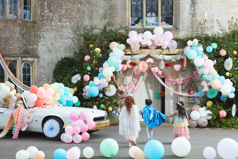 Colorful outdoor party setup with balloon garlands and a decorated car outside a stone building