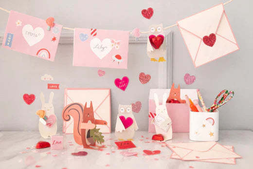 Valentine's envelopes in pink and white with hearts hang from a banner above Hugger crafts created in designs of woodland animals such as squirrels and bunnies. Glitter heart stickers and felt heart hair slides are also placed amongst the cards.