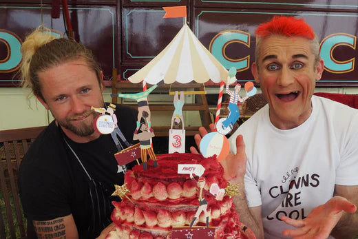 Two members of Gifford's Circus show off a red cake topped with circus characters and a big top, including strong men, acrobats, and clowns.