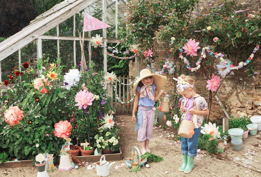 Two children in sun hats explore a colorful flower garden beside a greenhouse with bunting.