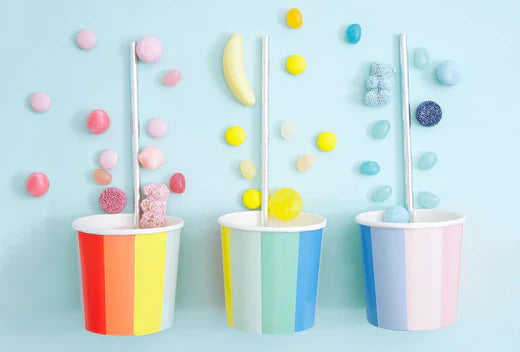 Three pastel rainbow cups with straws sit on a light blue background, surrounded by colorful candies.