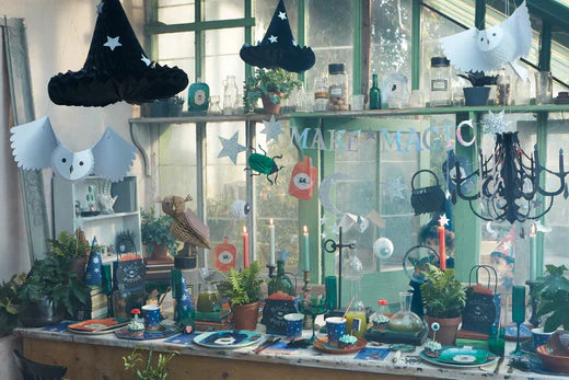 A greenhouse decorated with Magic Potion partyware, including napkins and plates, with honeycomb witches' hats and owls hanging from the ceiling above a garland featuring cauldrons and bugs.