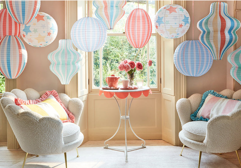 Pastel party nook with striped paper lanterns hanging from the ceiling, two plush cream chairs, and a round table set with a pink teapot and flowers.