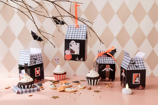 Various Halloween treat boxes are displayed on a table with black gingham tops and Halloween characters such as ghosts, black cats, and witches on the black sides of the boxes. Halloween cookies are placed beside the boxes.