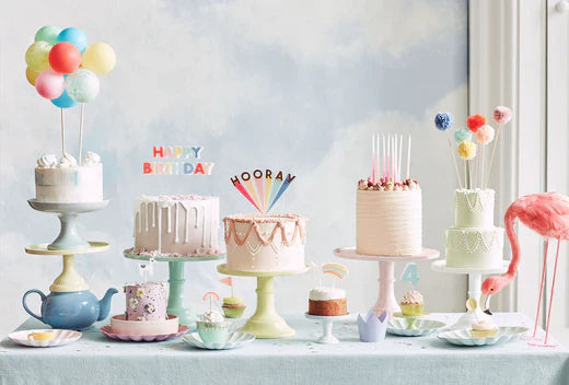 A line of large cakes covered in frosting placed on cake stands display a range of colourful candles and cake toppers.