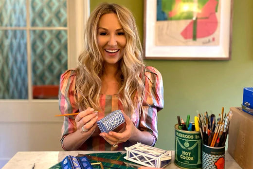 Lucy Clayton smiles at the camera whilst holding a paintbrush and trinket box, with other craft supplies on the table in front of her.