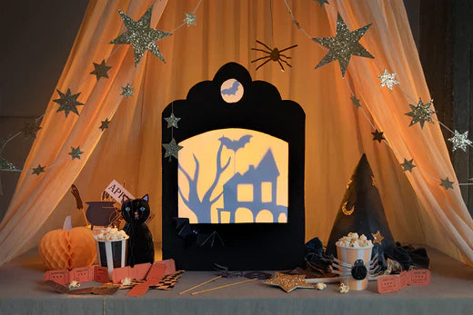 A small cardboard shadow theatre with Halloween-inspired shadows sits under a star garland and orange tulle, and is surrounded by honeycomb pumpkins and cat, a witch's hat, and striped cups with popcorn.
