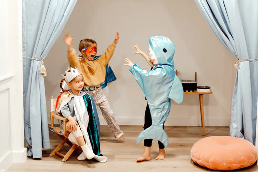 3 young children wear kids' costumes; from left to right, each wear a rainbow costume and crown, a superhero costume, and shark outfit.