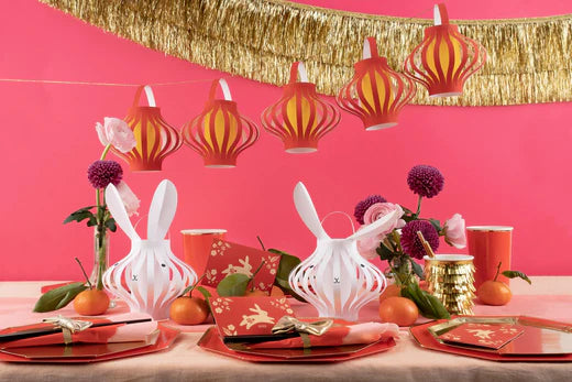 Party supplies inspired by the Lunar New Year are laid on a table, including red plates, bunny lanterns, and red and gold napkins. A gold tinsel banner and red lantern garland hang from the ceiling.