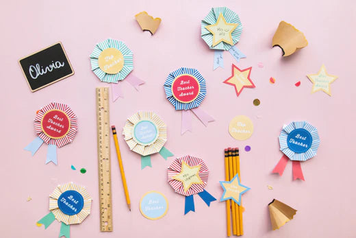 Paper rosettes in a mix of yellow, blue, and red that read 'Best Teacher' are featured against a pink background.
