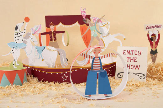 Cardboard circus characters surround a stage, including clowns, horses, acrobats, and a dog balancing a red ball on its nose.