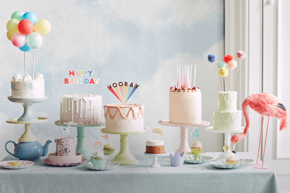 A row of cakes and cupcakes display a variety of candles, such as long glitter candles and statement cake toppers, and smaller designs including rainbows and numbers.