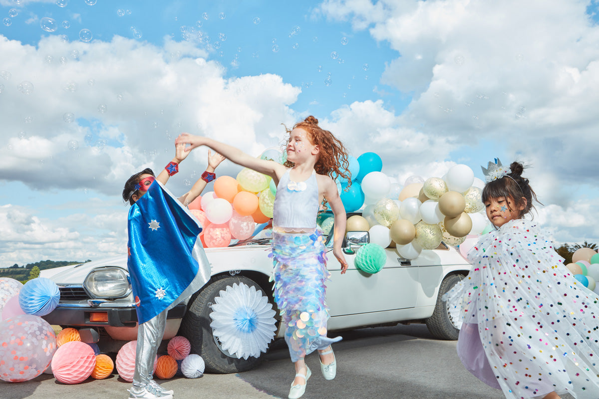 3 children wearing costumes, from left to right: a superhero, mermaid, and spotty cape stand amongst balloon arches and honeycomb ornaments.