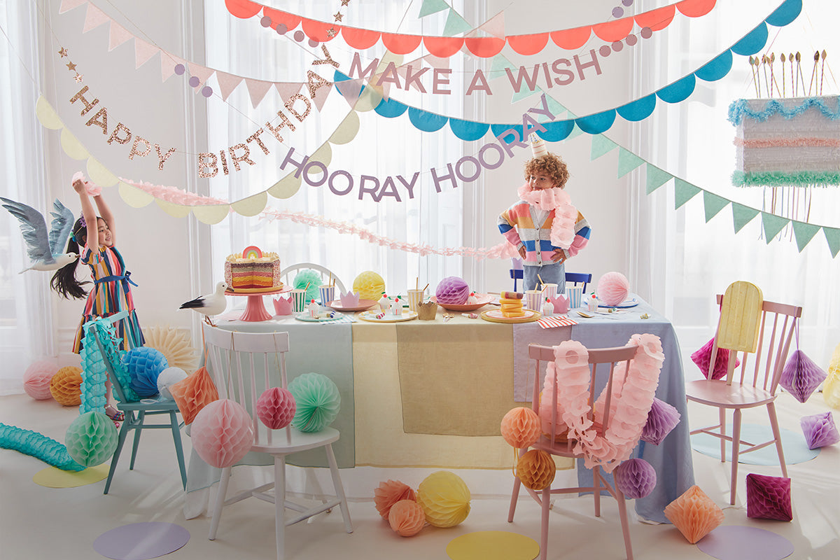 A little girl and boy standing around a colorful table lined with honeycomb decorations, bright garlands and banners, and rainbow tableware