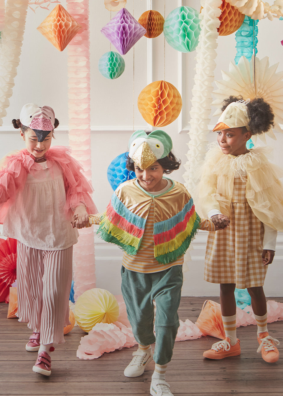 3 children dressed in various animal costumes, including a flamingo and octopus, standing in a room filled with pastel honeycomb hanging decorations and garlands