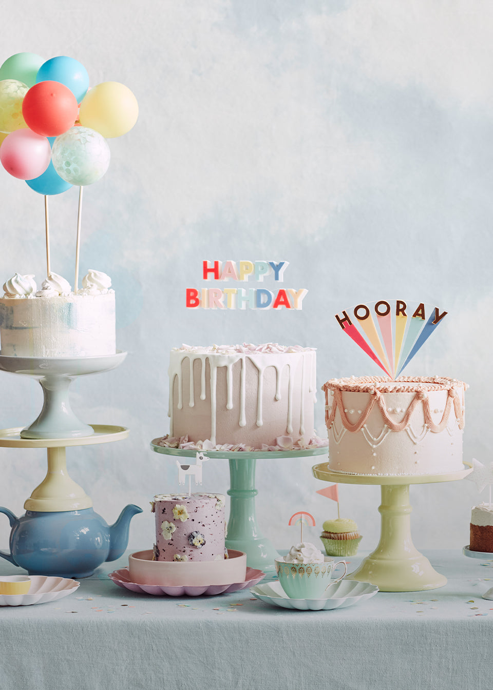 A row of cakes and cupcakes display a variety of candles, such as 'Happy Birthday' candles and statement cake toppers, and smaller designs including rainbows and animals.