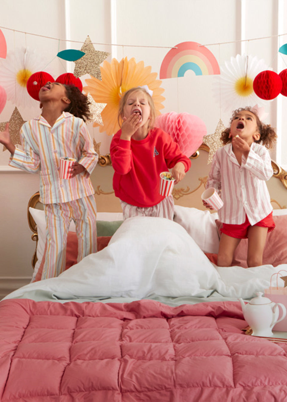 3 young girls stand in their pyjamas atop a bed and eat sweets whilst a pastel honeycomb garland with rainbows and hearts stretches across the wall in the background.