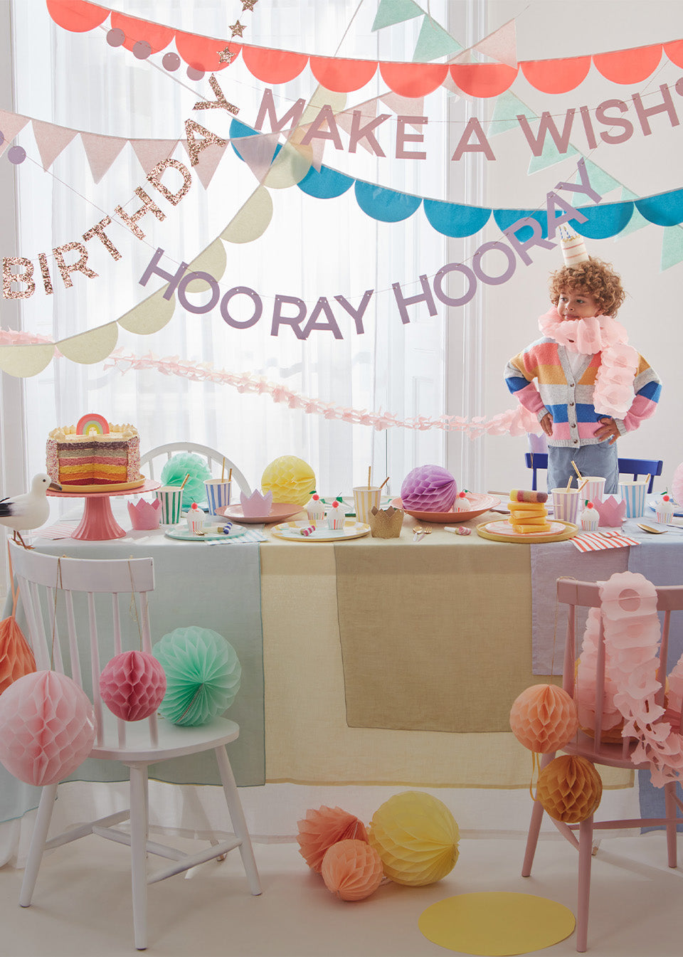 A little boy is standing around a colorful table lined with honeycomb decorations, bright garlands and banners, and rainbow tableware