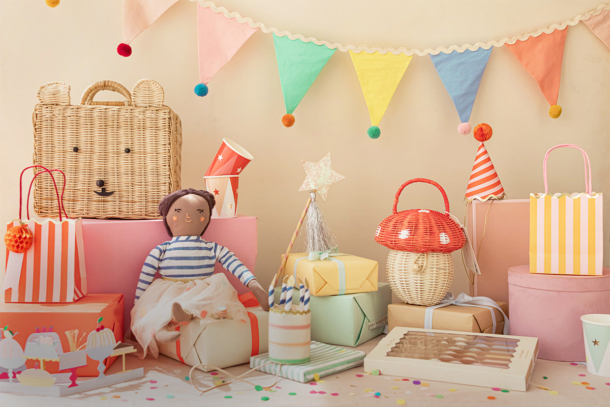 Amid rainbow confetti, a variety of gifts, including a doll, table candles, baskets, gift bags, and birthday partyware.