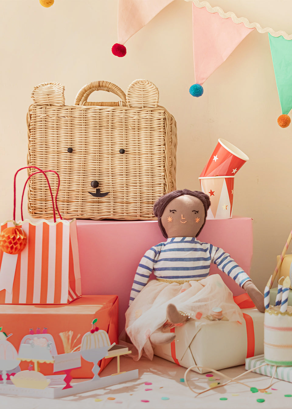 Amid rainbow confetti, a variety of gifts, including a doll, basket, gift bag, and birthday partyware.