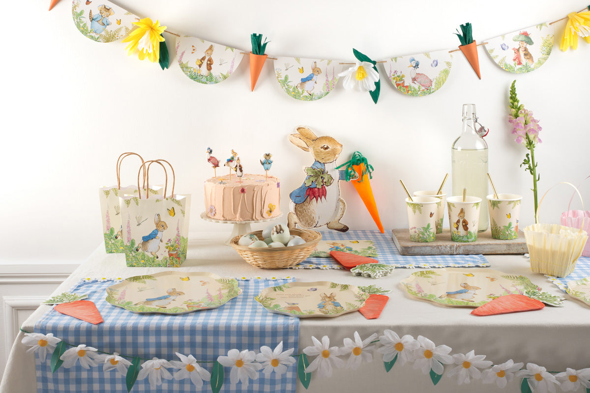 Peter Rabbit decorations are placed across a blue gingham tablecloth, including Peter Rabbit plates, garlands, cups, party bags, and napkins.