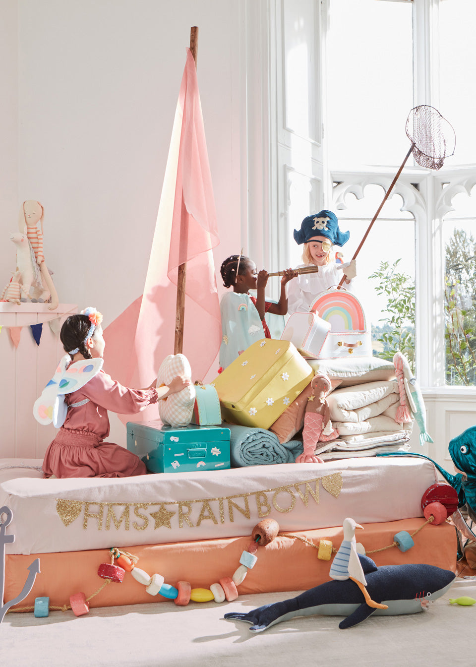 3 young kids wearing costumes play in a pillow fort decorated with colorful soft toys, such as rabbits, mermaid dolls, and a shark, and other accessories.